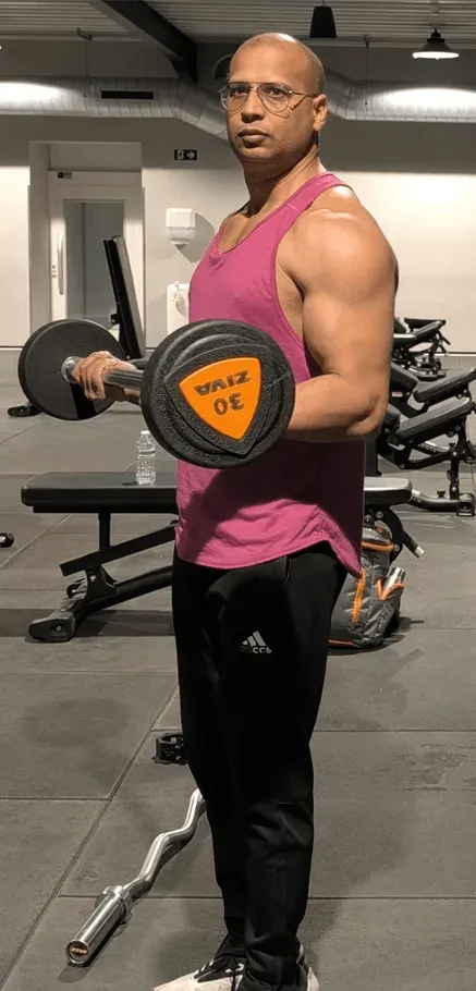 Ashish doet een curl met een beladen halterstang, staand in de fitnesszaal