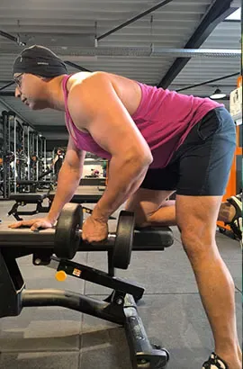 Ashish doet een roeibeweging met een dumbbell, met één knie en één hand op een bank